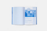 Open publication 'Ministry of Loneliness', the page to the left is white and the page to the right contains an image of an interior space in a rectangle shape in a blue colour and there is a series of text below the image. The image of the publication is taken on a white background.