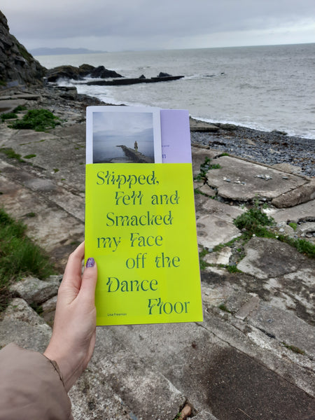 A hand holds the publication Slipped, Fell and Smacked my Face of the Dance Floor up. The book had  bright green and purple cover. In the backgound is a rocky shore. 
