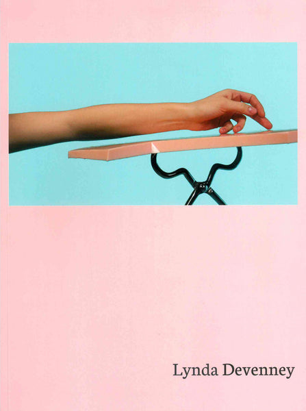 A pastoral pink cover with the authors name ' Lynda Devenney' written in black serif font in the bottom right corner. The top half of the cover has the image of a forearm and hand resting on a plank-type scultpure, the background is a bright pastel blue.